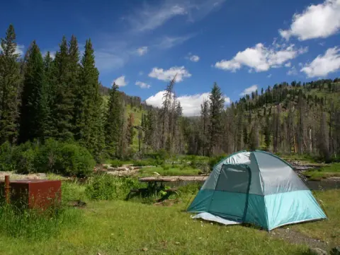 A bright blue-green tent with Slough Creek and woods behind