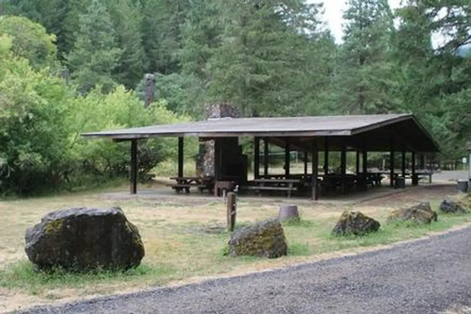 Large picnic shelter with stone fireplace and chimney and boulders lining a gravel road in front of fir forest and shrubs.