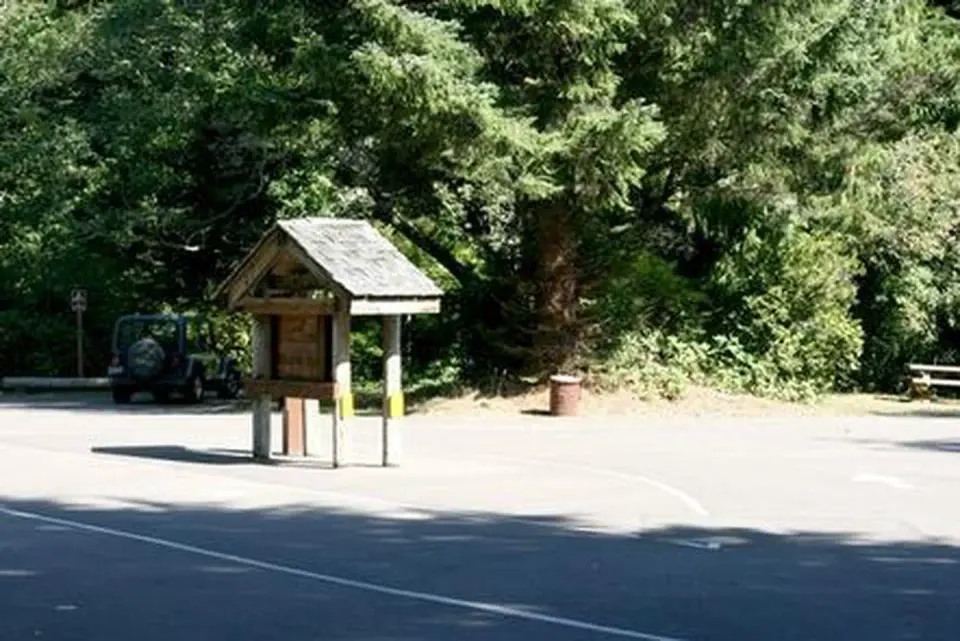 Paved entry with kiosk and fee tube in front of parking area and large evergreen shrub tree.