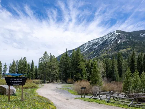 Oh Be Joyful Campground sign, gravel road, snow dusted mountain, forest, yellow wildflowers